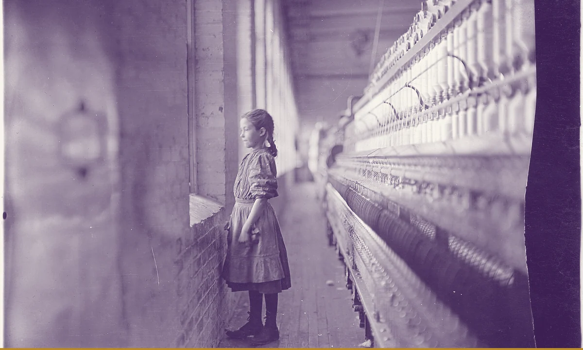 A young girl standing at a spinning frame in a cotton mill, looking out of a window.